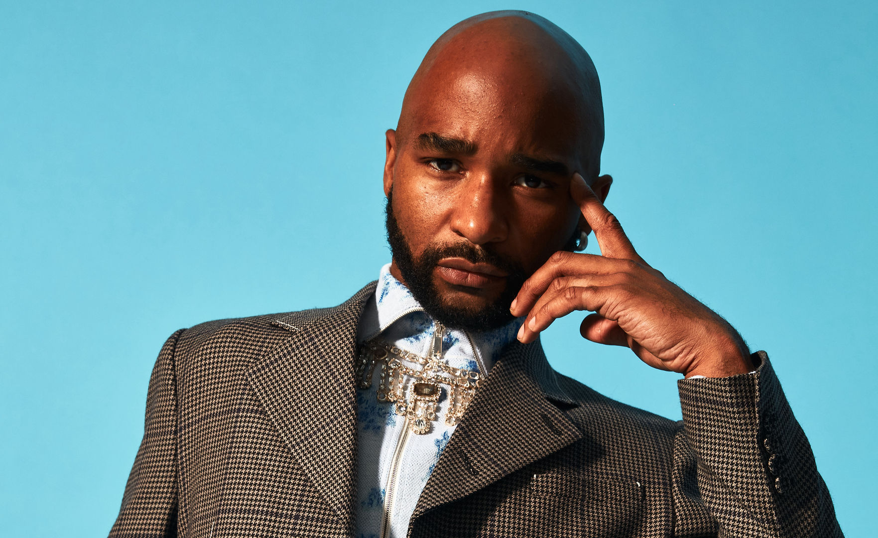 A headshot of artist Jacolby Satterwhite, wearing a gray jacket, light blue button-down shirt buttoned to the top, and a pendant around his neck.